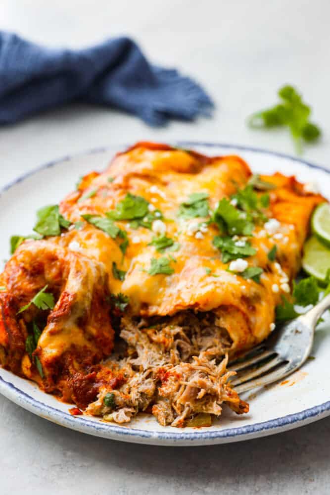 These delicious enchiladas are filled with juicy pork carnitas and smothered in melted cheese. Topped off with homemade red enchilada sauce, this is a dinner that the whole family will go crazy over. Closeup of pork enchiladas with a bite taken out of them.