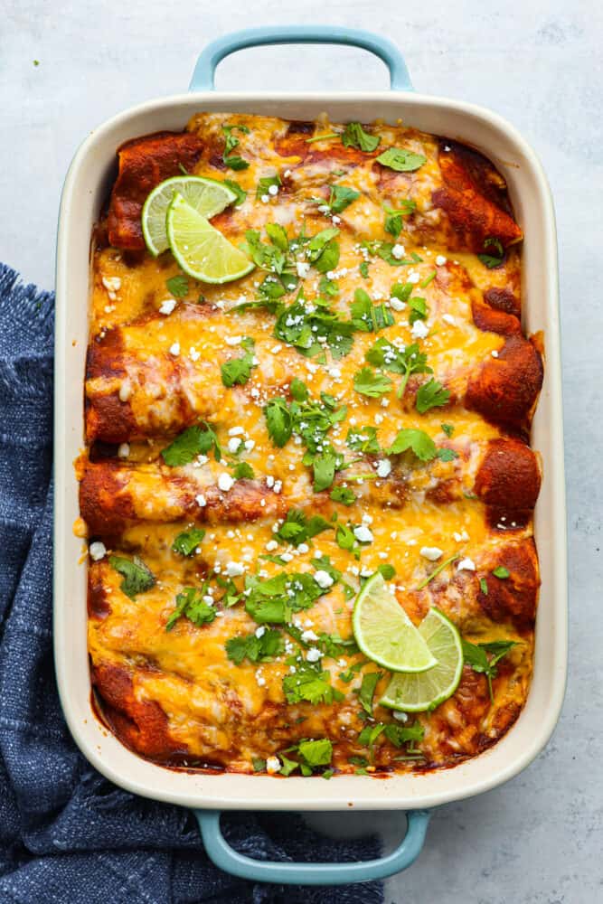 These delicious enchiladas are filled with juicy pork carnitas and smothered in melted cheese. Topped off with homemade red enchilada sauce, this is a dinner that the whole family will go crazy over. A blue and white casserole dish filled with baked enchiladas.