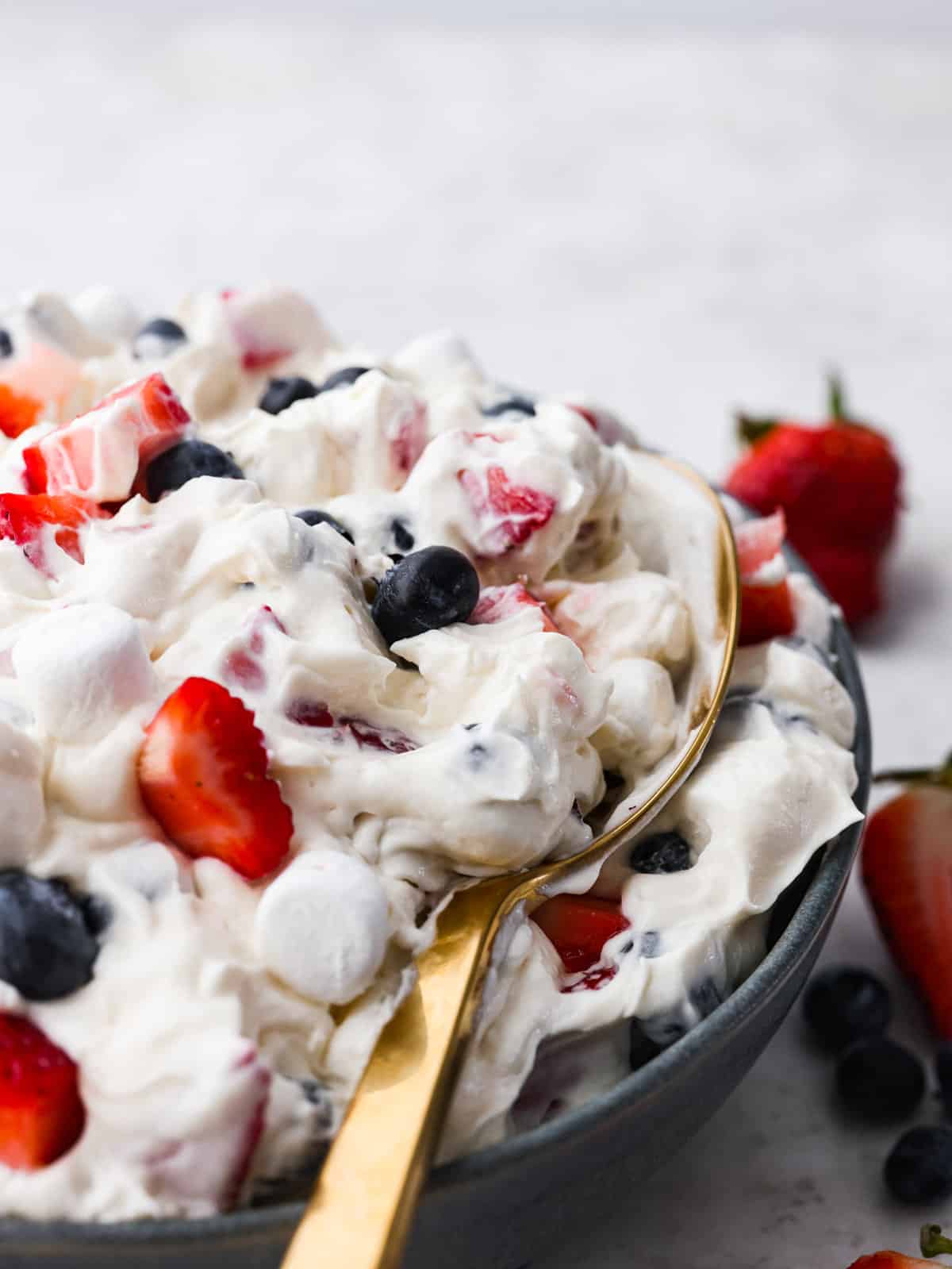 Introducing the red, white, and blue cheesecake salad, a delightful and patriotic summer fruit salad that combines the vibrant flavors of strawberries and blueberries with creamy cheesecake and fluffy marshmallows. It's super easy to throw together and a hit at every potluck! Cheesecake salad being scooped up with a gold spoon.