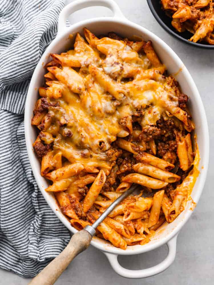 Swap out the bun for this sloppy joe casserole full of pasta and a sweet ground beef mixture. All of the meaty and delicious flavors of a sloppy joe sandwich transform into an easy weeknight meal your family will love! Top view of sloppy Joe casserole in a white baking dish. A striped blue and white towel and black bowl filled with casserole are next to the baking dish.