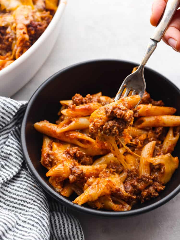 Swap out the bun for this sloppy joe casserole full of pasta and a sweet ground beef mixture. All of the meaty and delicious flavors of a sloppy joe sandwich transform into an easy weeknight meal your family will love! Close view of a fork lifting a bite of the casserole out of a black bowl. A striped blue and white towel along with the casserole dish are next to the serving bowl.