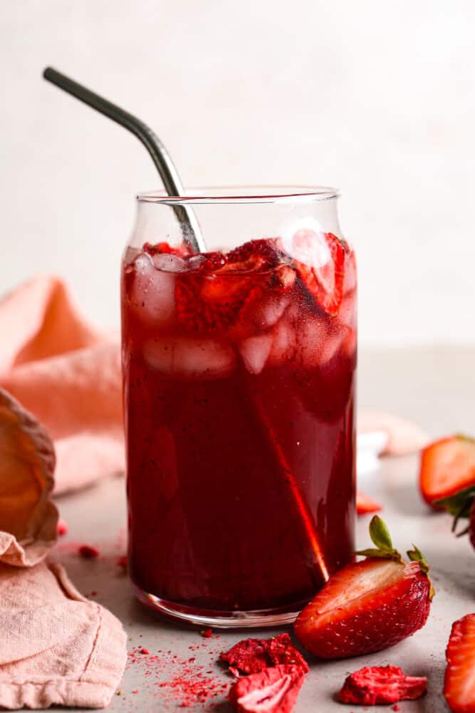 This Strawberry Acai Refresher is a refreshing drink that's sure to help you beat the heat! Whether you're lounging by the pool, hosting a summer soirรฉe, or simply in need of a tropical escape, this refresher will be your go-to beverage. The side view of a strawberry acai refresher in a clear cup with a metal straw and strawberries scattered around the cup.