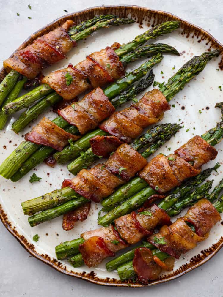 Bacon wrapped asparagus is a smoky and savory side dish that will be a staple for mealtime. Each bundle is glazed with maple syrup for caramelized flavor you wonโt be able to get enough of! Top-down view of bacon wrapped asparagus on a white and brown plate.