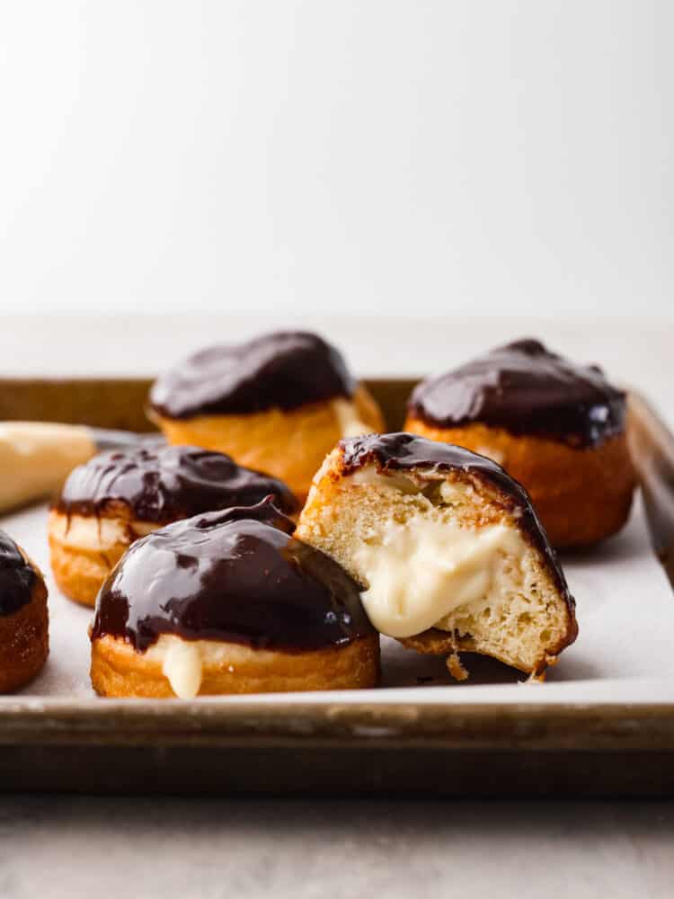 Treat yourself to a batch of homemade Boston Cream Donuts covered in chocolate ganache. These delightful treats are made from scratch and create an irresistible combination of fluffy donuts, creamy filling, and delicious chocolate topping. The side view of a Boston cream donut cut open with the filling coming out.