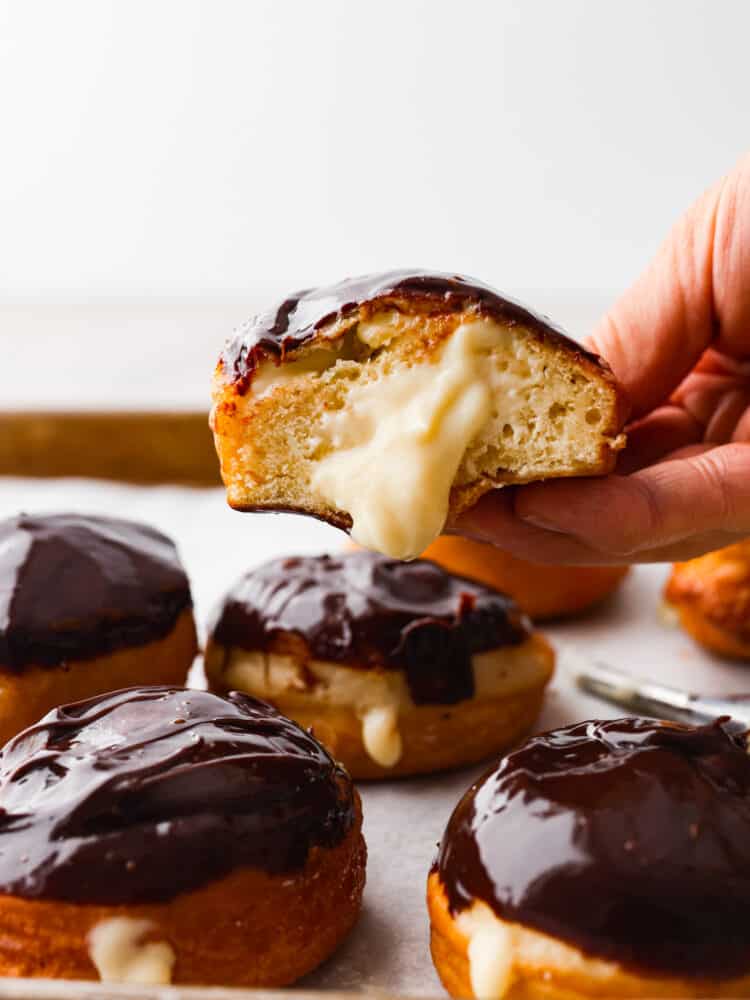 Treat yourself to a batch of homemade Boston Cream Donuts covered in chocolate ganache. These delightful treats are made from scratch and create an irresistible combination of fluffy donuts, creamy filling, and delicious chocolate topping. Someone holding a Boston cream donut that is cut in half.
