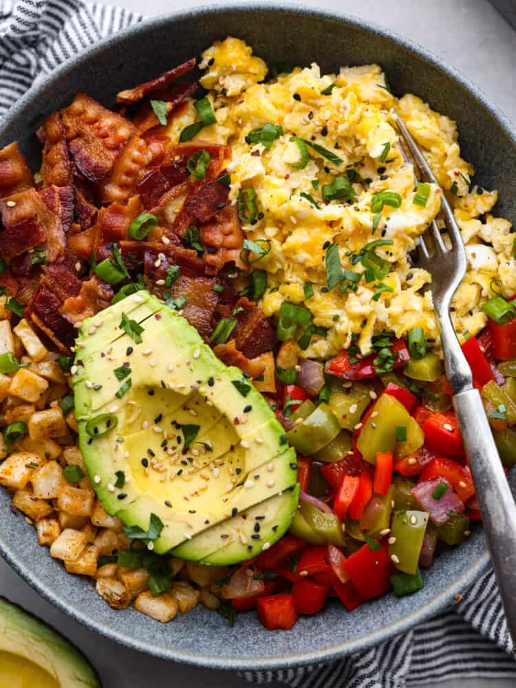 All your favorite breakfast ingredients together in one hearty and delicious breakfast bowl! Full of eggs, bacon, potatoes, and veggies, this bowl will keep you satisfied and fueled all morning long. Close view of a breakfast bowl with a fork in the bowl. Eggs, cheese, bacon, potatoes, and vegetables are in the bowl with chopped cilantro on top.