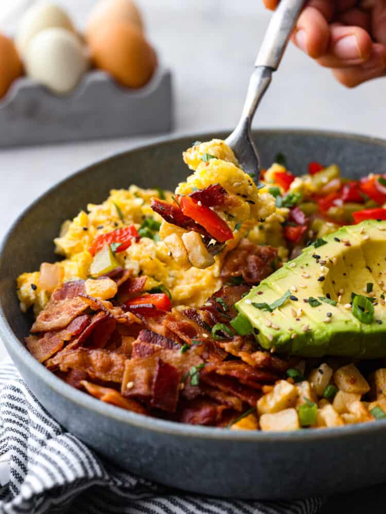All your favorite breakfast ingredients together in one hearty and delicious breakfast bowl! Full of eggs, bacon, potatoes, and veggies, this bowl will keep you satisfied and fueled all morning long. Close view of a fork lifting up eggs, bacon, and veggies from the breakfast bowl.