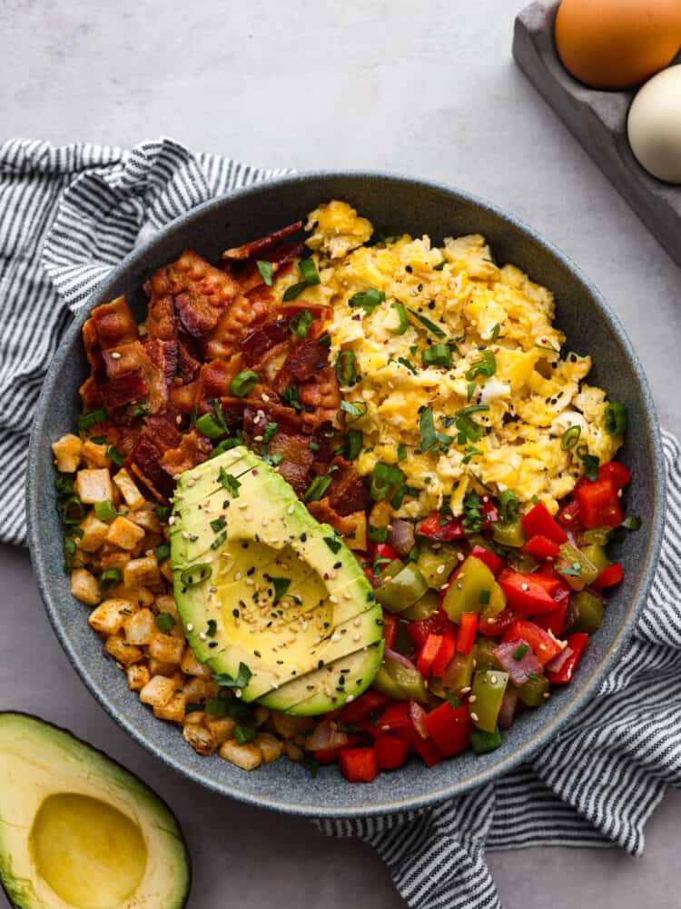All your favorite breakfast ingredients together in one hearty and delicious breakfast bowl! Full of eggs, bacon, potatoes, and veggies, this bowl will keep you satisfied and fueled all morning long. Top view of a breakfast bowl with sliced avocado on top.