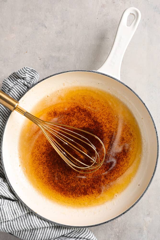 Learning how to brown butter will be your secret weapon in the kitchen! Easily done over the stove, butter slowly melts and releases a nutty toasted flavor that will enhance any sweet or savory recipe. Top view of browned butter in a white skillet. A gold whisk is inside the skillet with a blue and white striped towel next to the skillet.