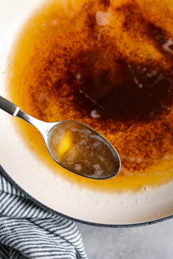 Learning how to brown butter will be your secret weapon in the kitchen! Easily done over the stove, butter slowly melts and releases a nutty toasted flavor that will enhance any sweet or savory recipe. Close up view of a spoon lifting up brown butter out of a white skillet. A blue and white striped towel is next to the skillet.
