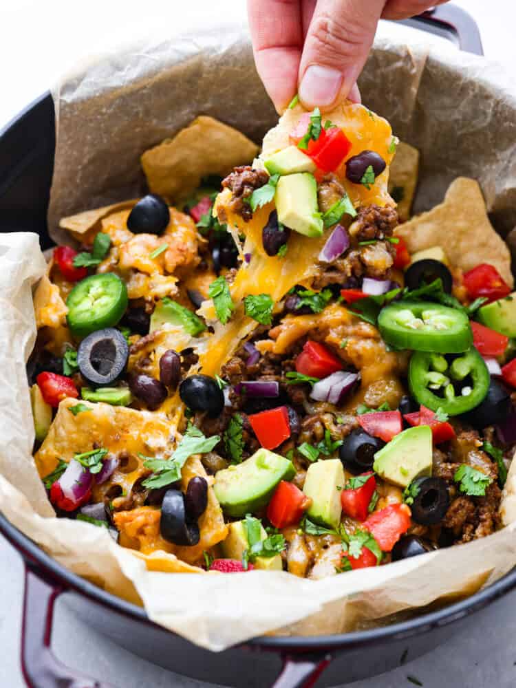 These campfire nachos are the ultimate outdoor comfort food! Packed with layers of seasoned ground beef, refried beans, and gooey cheese, these nachos are then topped with your favorite veggies and will be a new summertime favorite! A hand reaching into the pan and taking out a chip.