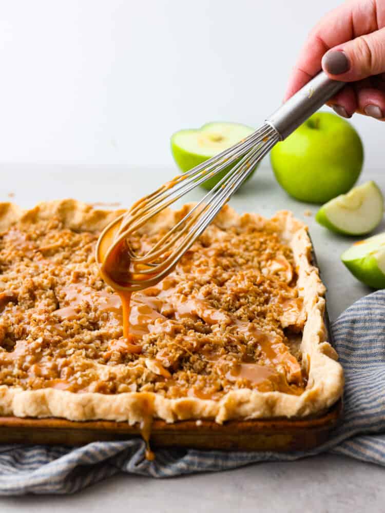 Caramel Apple Slab Pie has a thin and flaky melt-in-your-mouth crust that is topped with juicy apples and a crisp topping. It is drizzled in homemade caramel and perfect for feeding a crowd! A whisk drizzling caramel onto the slab pie.