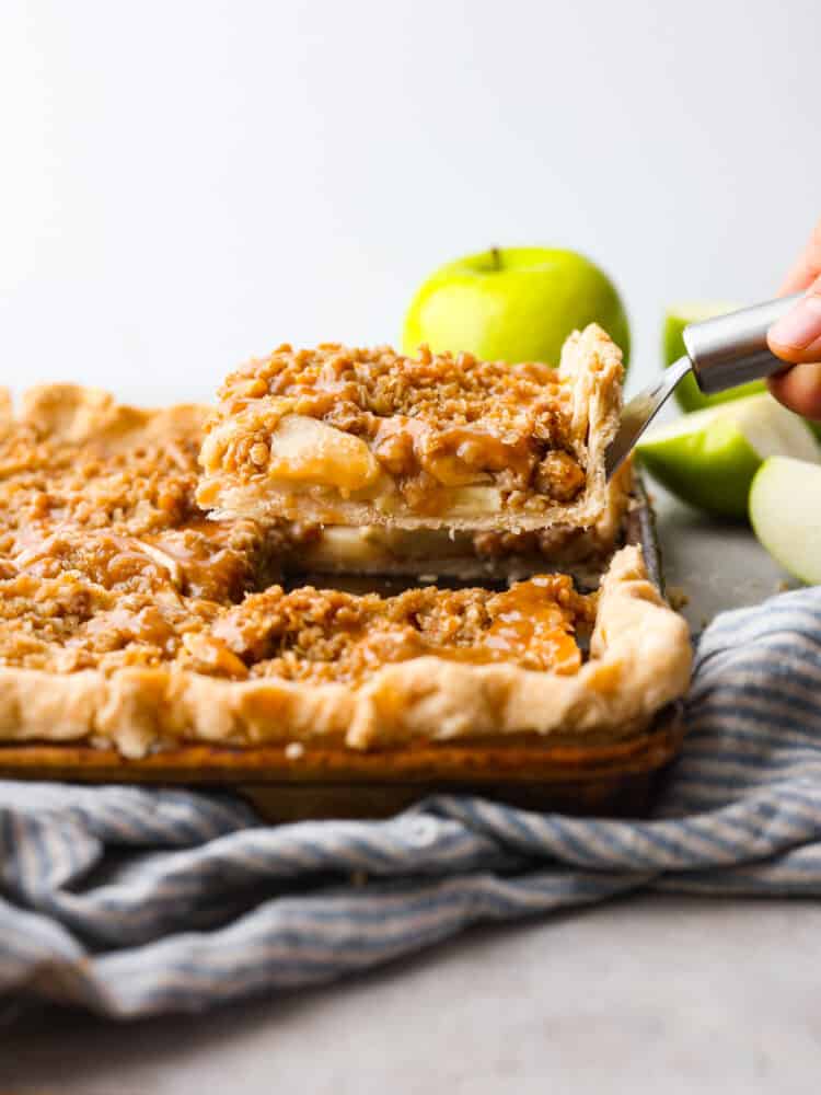 Caramel Apple Slab Pie has a thin and flaky melt-in-your-mouth crust that is topped with juicy apples and a crisp topping. It is drizzled in homemade caramel and perfect for feeding a crowd! the side view of a slice of caramel apple slab pie being served.