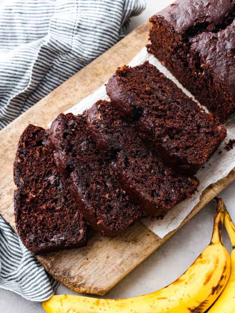 Chocolate banana bread is a sweet and decadent banana bread made for chocolate lovers! Extra moist from the ripe bananas, and loaded with rich chocolate flavors from the cocoa powder and chocolate chips. Top view of sliced chocolate banana bread on a wood cutting board. Bananas and a striped towel are next to the cutting board.