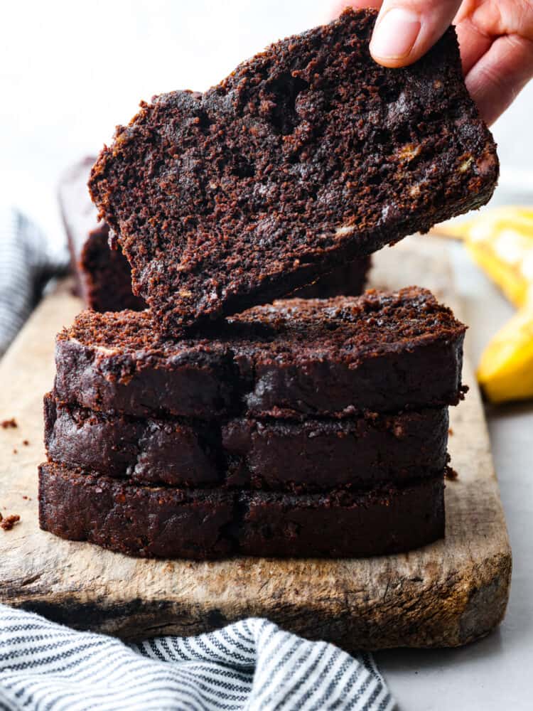 Chocolate banana bread is a sweet and decadent banana bread made for chocolate lovers! Extra moist from the ripe bananas, and loaded with rich chocolate flavors from the cocoa powder and chocolate chips. Side view of stacked slices of chocolate banana bread. A hand is lifting a slice of bread.