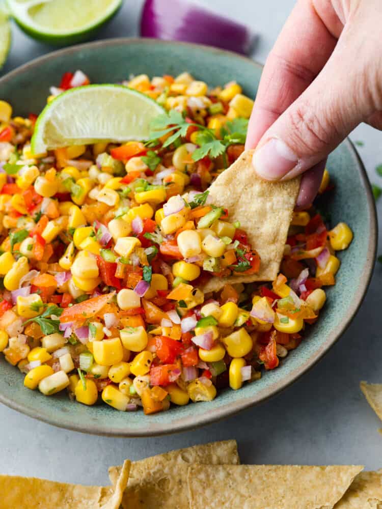 Experience the ultimate freshness and flavor with this easy Corn Salsa recipe. Packed with fresh ingredients like red onion, jalapeรฑo, and lime juice, it's the perfect blend of flavors for a refreshing summer appetizer.. A hand dipping a tortilla chip into the corn salsa.