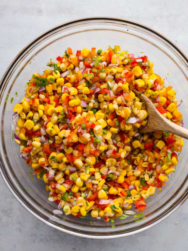 Experience the ultimate freshness and flavor with this easy Corn Salsa recipe. Packed with fresh ingredients like red onion, jalapeรฑo, and lime juice, it's the perfect blend of flavors for a refreshing summer appetizer. The top view of the ingredients being mixed with a wooden spoon.