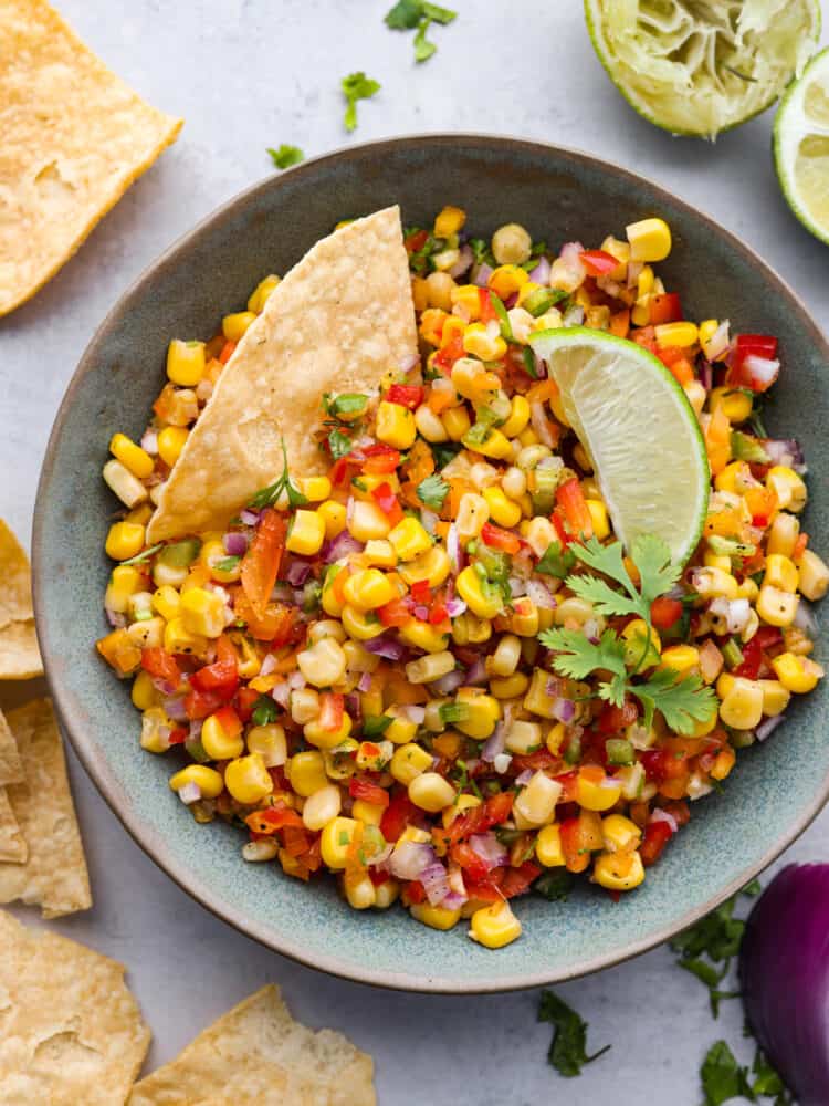 Experience the ultimate freshness and flavor with this easy Corn Salsa recipe. Packed with fresh ingredients like red onion, jalapeรฑo, and lime juice, it's the perfect blend of flavors for a refreshing summer appetizer. The top view of a bowl of corn salsa garnished with cilantro and topped with a lime and a tortilla chip.
