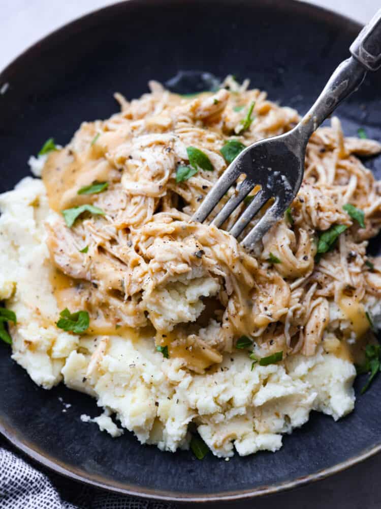 Whip up a comforting meal with this delicious Crockpot Chicken and Gravy recipe. Cooked to perfection in the slow cooker, the result is tender, juicy chicken smothered in a rich gravy, making it the perfect companion to rice or potatoes for a hearty and satisfying dinner! A close up shot of chicken and gravy on a pile of mashed potatoes on a black plate with a silver fork.