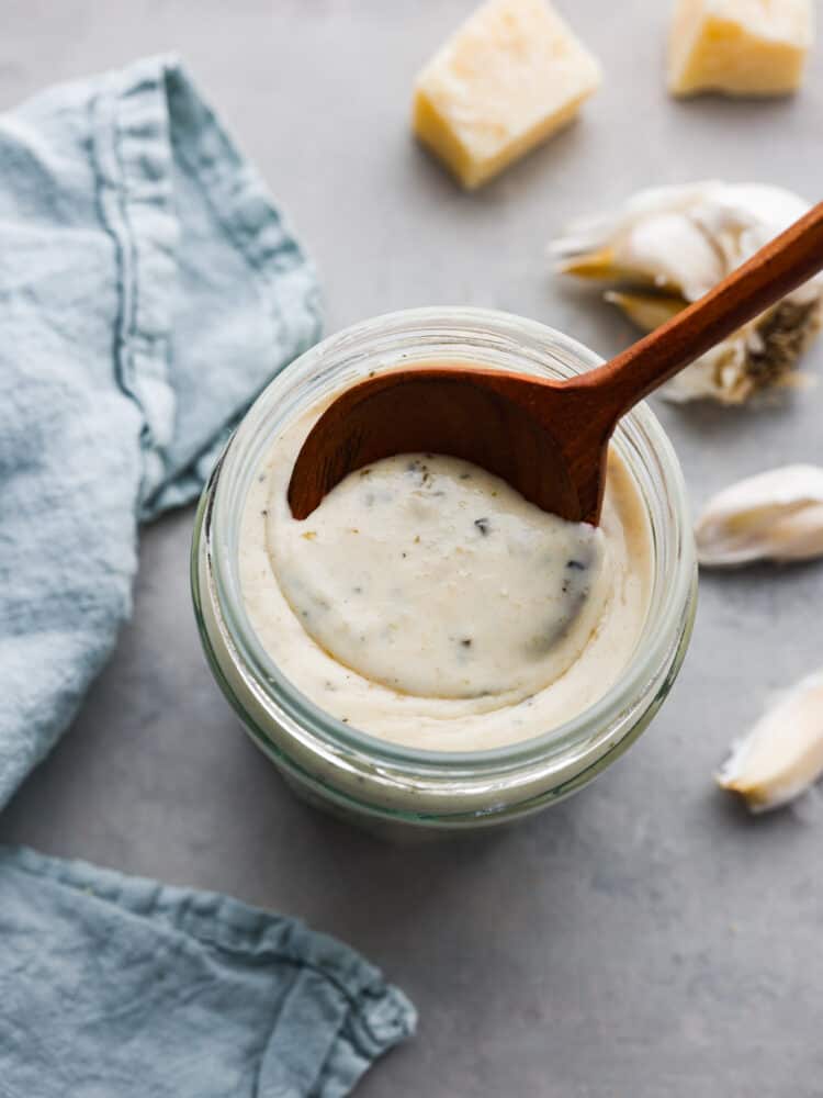 Garlic parmesan sauce is a copycat recipe from the famous Buffalo Wild Wings restaurant. This creamy garlic sauce is best known as a wing sauce, but it's perfect for pasta, on pizza, or used as a dipping sauce. Close up view garlic parmesan sauce in a glass jar with a wood spoon in the jar. Fresh garlic and parmesan cheese next to the jar.