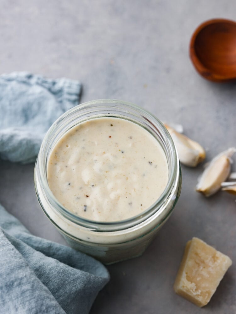Garlic parmesan sauce is a copycat recipe from the famous Buffalo Wild Wings restaurant. This creamy garlic sauce is best known as a wing sauce, but it's perfect for pasta, on pizza, or used as a dipping sauce. Close up view garlic parmesan sauce in a glass jar. Fresh garlic and parmesan cheese next to the jar. A light blue towel is styled next to the jar.