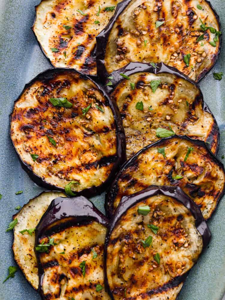 Grilled eggplant has a perfect golden char on the outside and is deliciously seasoned and tender on the inside. Discover how simple and impressive grilled eggplant is when served as a side dish to any meal. Close up view of grilled eggplant on a blue platter garnished with chopped parsley.