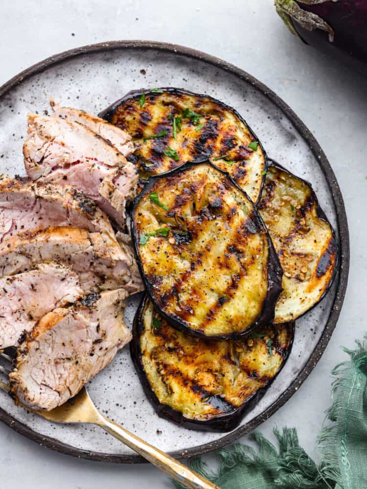Grilled eggplant has a perfect golden char on the outside and is deliciously seasoned and tender on the inside. Discover how simple and impressive grilled eggplant is when served as a side dish to any meal. Top view of grilled eggplant on a white plate along with sliced pork and gold fork.