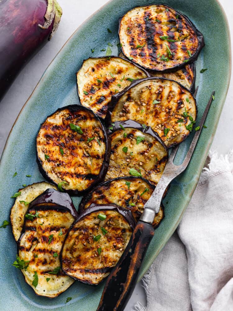 Grilled eggplant has a perfect golden char on the outside and is deliciously seasoned and tender on the inside. Discover how simple and impressive grilled eggplant is when served as a side dish to any meal. Top view of grilled eggplant on a blue oval shaped platter with a serving fork is on the platter.