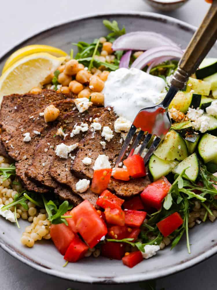 Take lunchtime up a notch with this incredible gyro bowl! It has tender gyro meat, fluffy couscous, crunchy veggies, and zesty tzatziki all in one bowl. Healthy, hearty, and totally irresistible. Taking a bite of gyro meat with a fork.