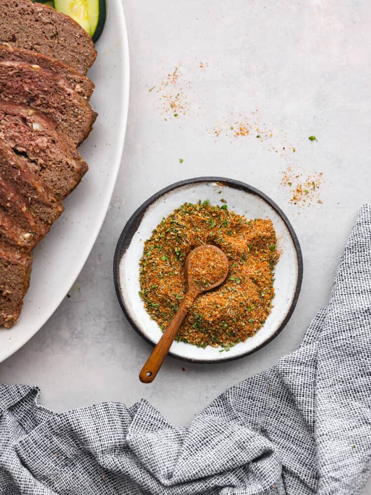 Gyro seasoning is a Greek spice blend made with a delicious combination of spices and herbs. Enhance your chicken, beef, lamb, or pork with the incredible Mediterranean flavors that gyros are known for! Top view of gyro seasoning in a white bowl with a wood spoon. A platter of gyro meat and a kitchen towel are next to the seasoning.