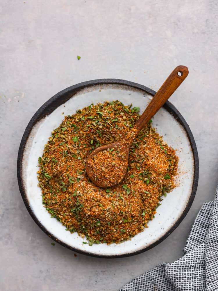 Gyro seasoning is a Greek spice blend made with a delicious combination of spices and herbs. Enhance your chicken, beef, lamb, or pork with the incredible Mediterranean flavors that gyros are known for! Overhead view of gyro seasoning in a white bowl and a wood spoon.