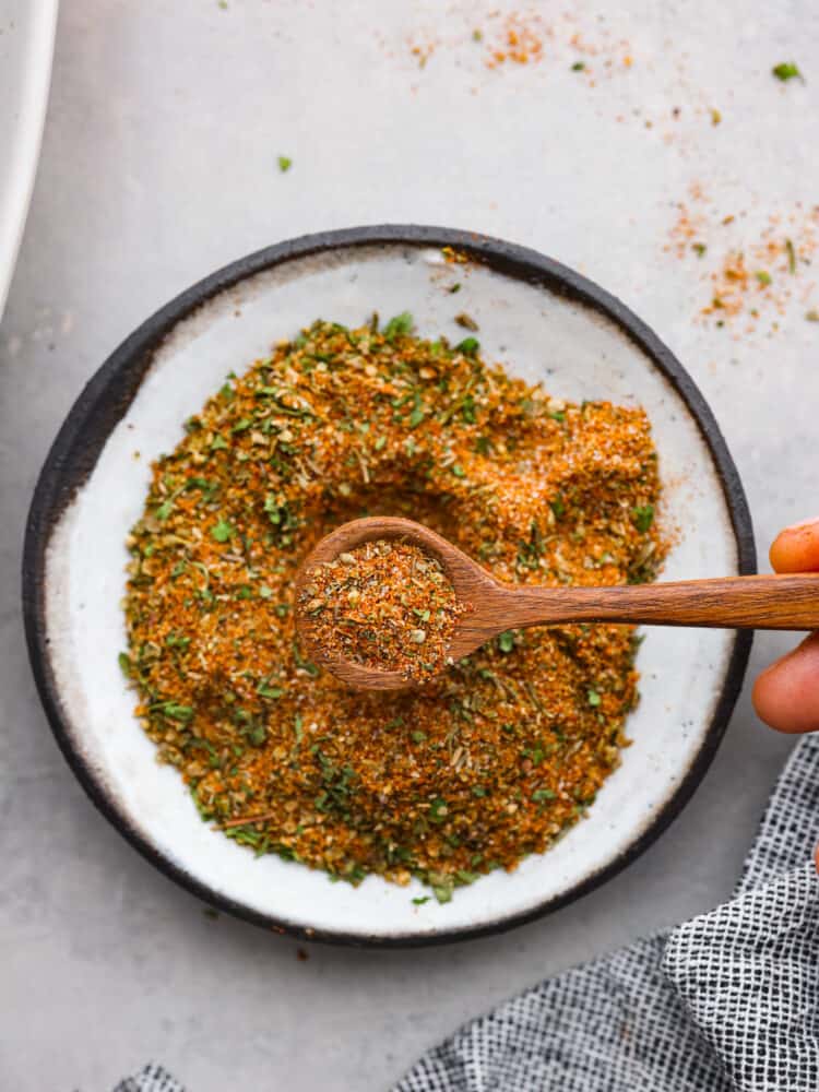Gyro seasoning is a Greek spice blend made with a delicious combination of spices and herbs. Enhance your chicken, beef, lamb, or pork with the incredible Mediterranean flavors that gyros are known for! Close up view of gyro seasoning in a white bowl with a wood spoon. The spoon is lifting seasoning out of the bowl.