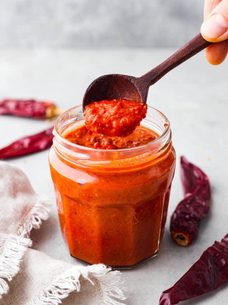Harissa paste is a fiery and delicious condiment from North Africa that packs a punch. It combines spicy dried guajillo peppers, smoky roasted red peppers, and other tasty seasonings to create a flavor explosion that can elevate any dish! Hero image of harissa paste in a glass jar.