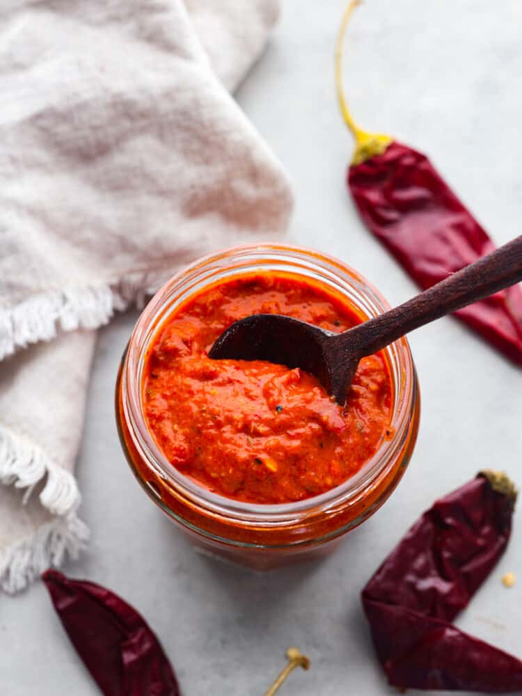 Harissa paste is a fiery and delicious condiment from North Africa that packs a punch. It combines spicy dried guajillo peppers, smoky roasted red peppers, and other tasty seasonings to create a flavor explosion that can elevate any dish! Top-down view of a jar of harissa paste with a wooden spoon inside.