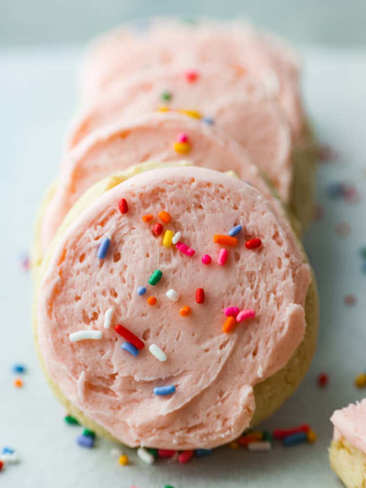 These classic Lofthouse Sugar Cookies are soft and puffy cake-like sugar cookies that taste just like the original! Topped with homemade buttercream frosting and rainbow sprinkles, these cookies will bring your right back to your childhood! Frosted lofthouse cookies in a line.