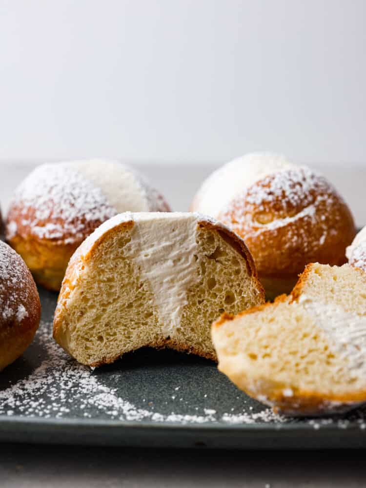 These airy, cream-filled pastries are called maritozzi and a must-make for any dessert lover. The buns have a soft, tender texture that pairs perfectly with the luscious vanilla filling! Top them off with a dusting of powdered sugar for a sweet Italian treat that everyone will love. Hero image of maritozzi buns. One is cut in half so the filling inside can be seen.