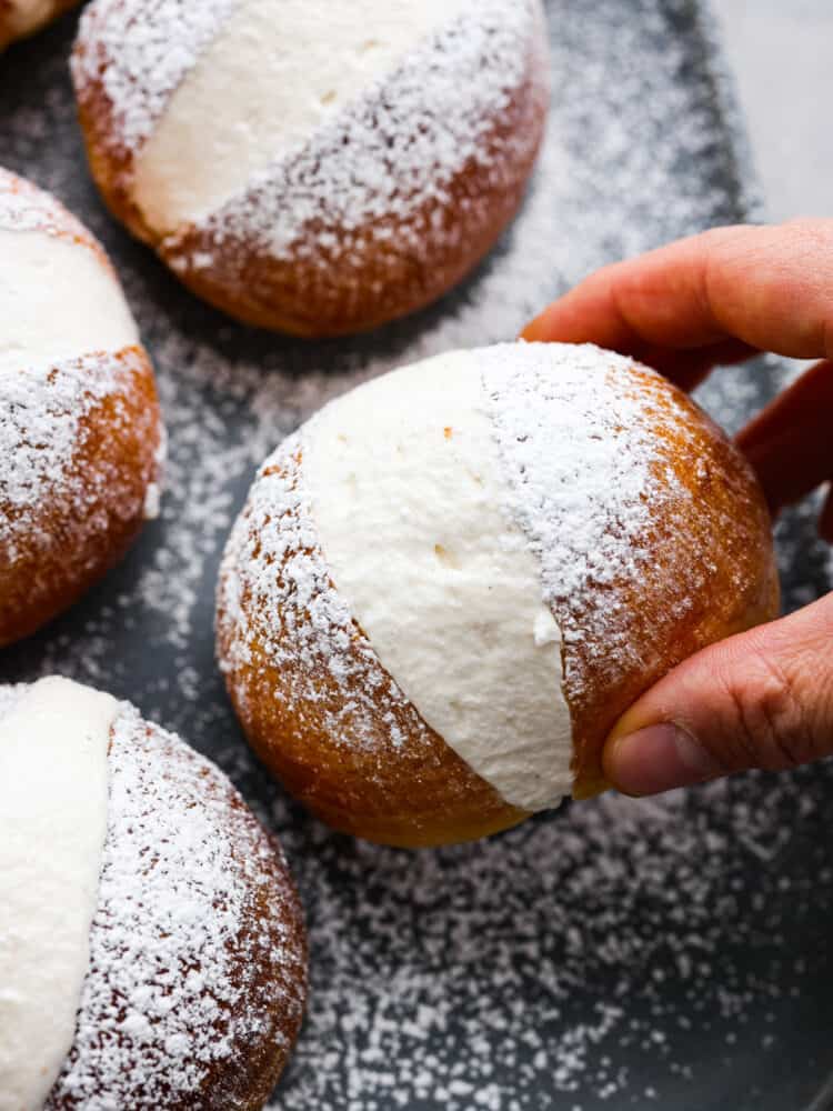 These airy, cream-filled pastries are called maritozzi and a must-make for any dessert lover. The buns have a soft, tender texture that pairs perfectly with the luscious vanilla filling! Top them off with a dusting of powdered sugar for a sweet Italian treat that everyone will love. A bun being picked up.