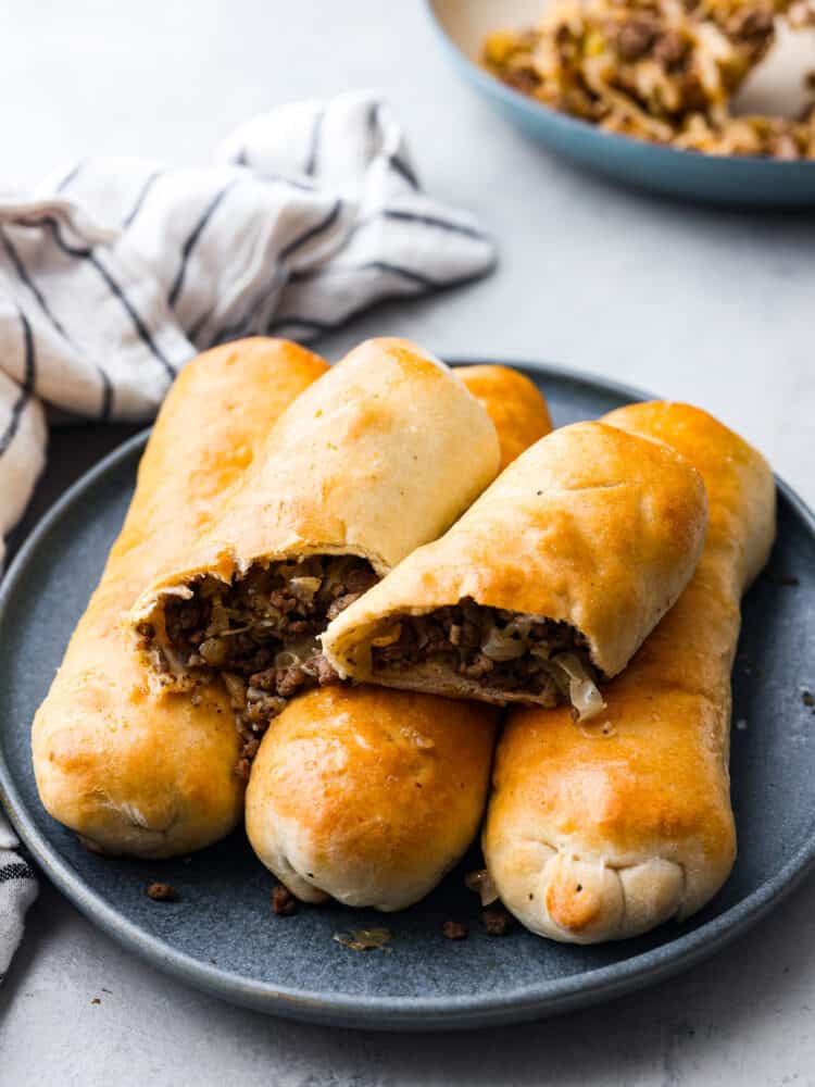 Runza is a tasty, handheld pastry stuffed with a scrumptious mix of ground beef, onions, garlic, cabbage, and spices, all snug inside soft pizza dough. Runzas on a gray plate. One is cut in half.