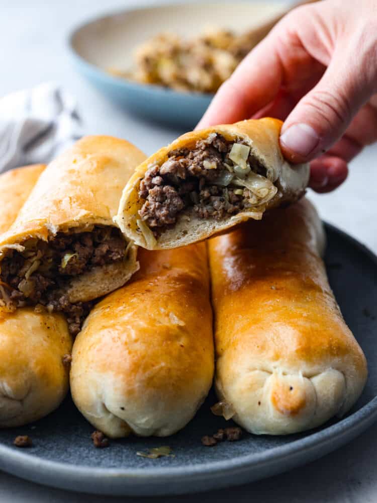 Runza is a tasty, handheld pastry stuffed with a scrumptious mix of ground beef, onions, garlic, cabbage, and spices, all snug inside soft pizza dough. Runzas stacked on top of each other. One is cut in half so you can see the filling.