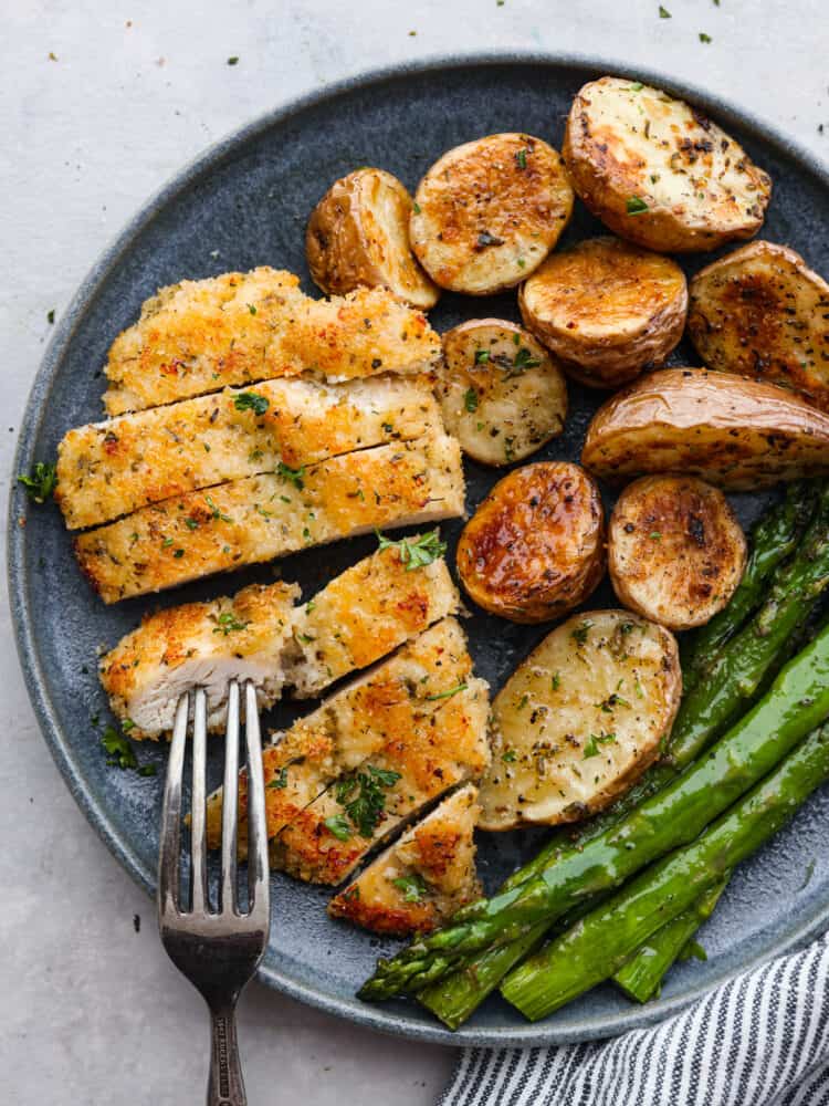Sheet Pan Crispy Parmesan Garlic Chicken is such an easy meal with crispy breaded chicken with potatoes and asparagus. It's easy to make, easy to clean up, and is the perfect meal for any occasion! Sliced chicken, potatoes and asparagus on a blue plate with a fork on top.