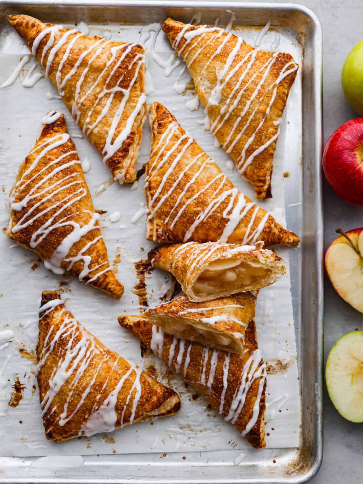 Treat yourself to delicious homemade apple turnovers with this simple recipe. These golden-brown puff pastry triangles are filled with canned apple pie, baked until flaky, and topped with a sweet powdered sugar glaze. The top view of apple turnovers on a baking sheet with parchment paper.