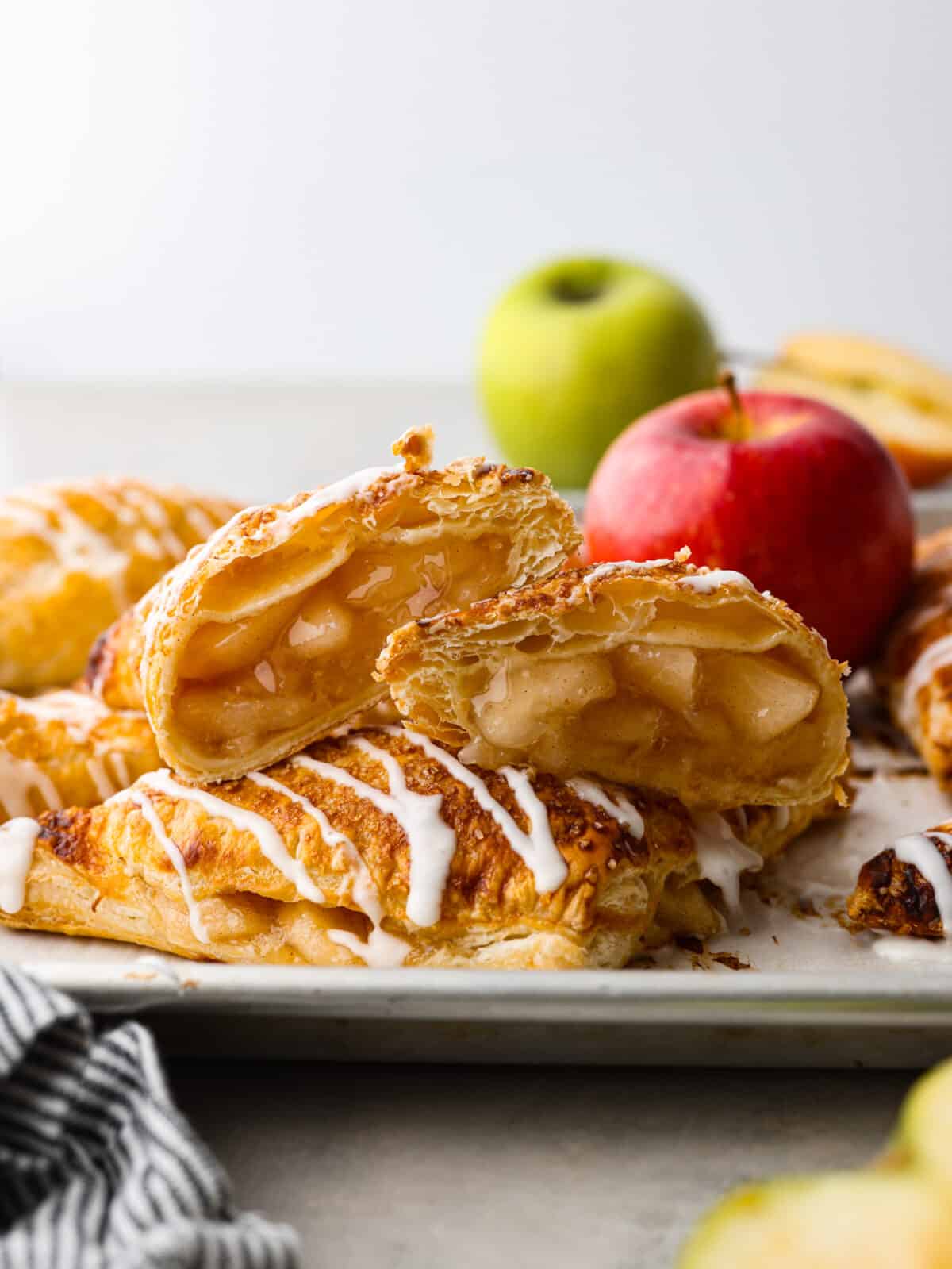 Treat yourself to delicious homemade apple turnovers with this simple recipe. These golden-brown puff pastry triangles are filled with canned apple pie, baked until flaky, and topped with a sweet powdered sugar glaze. 2 apple turnovers stacked on on another. the top one is cut open so that you can see the filling.
