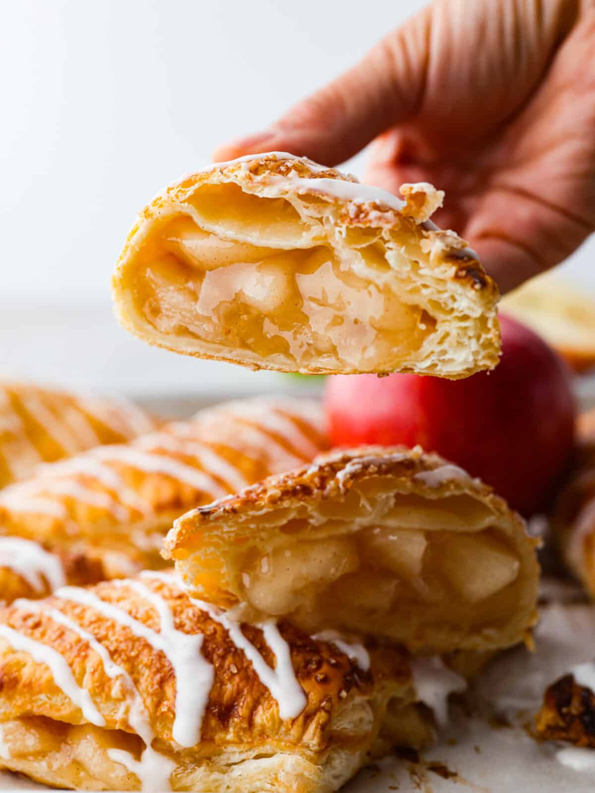 Treat yourself to delicious homemade apple turnovers with this simple recipe. These golden-brown puff pastry triangles are filled with canned apple pie, baked until flaky, and topped with a sweet powdered sugar glaze. A hand holding an apple turnover that is cut in half.