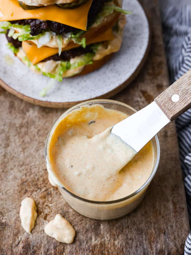 Level up your burgers with this tasty homemade Big Mac sauce! It's made with creamy mayo, pickle relish, vinegar, and a bunch of delicious seasonings. You'll always want it on hand! Hero image of Big Mac sauce in a glass container. There is a burger in the background.
