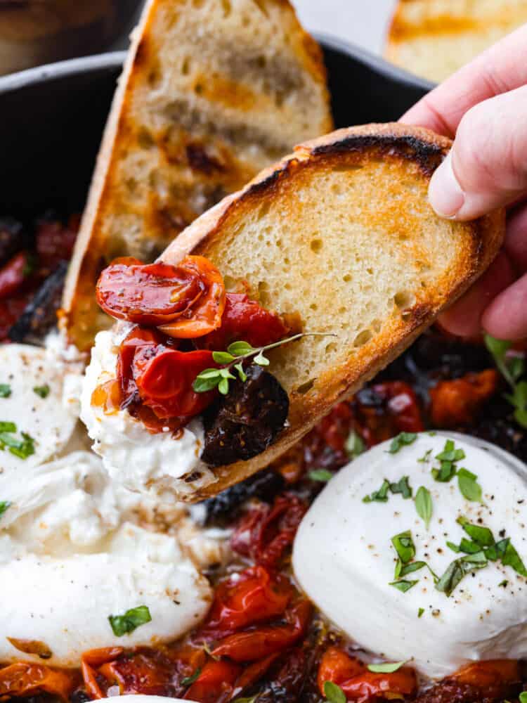 This burrata appetizer is a flavor bomb made with juicy cherry tomatoes, sweet dried figs, and a tangy balsamic kick. Topped with creamy burrata cheese, it's pure indulgence. Perfect for parties or whenever you want to treat yourself to something special! Scooping up tomatoes and cheese with a piece of toasted, sliced baguette.