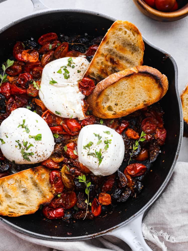 This burrata appetizer is a flavor bomb made with juicy cherry tomatoes, sweet dried figs, and a tangy balsamic kick. Topped with creamy burrata cheese, it's pure indulgence. Perfect for parties or whenever you want to treat yourself to something special! Top-down view of an appetizer made with cherry tomatoes, figs, and burrata cheese. There are 2 pieces of toasted baguette in the skillet.