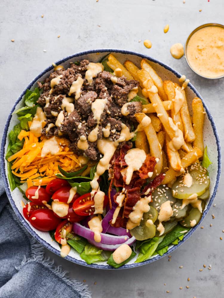 This deconstructed burger bowl is so hearty and satisfying, you'll love every bite! It features crispy fries, tender beef, pickles, and other fresh veggies all coated in homemade Big Mac sauce! Top-down view of a burger bowl made with beef, bacon, veggies, and shredded cheese, all topped with a creamy sauce.