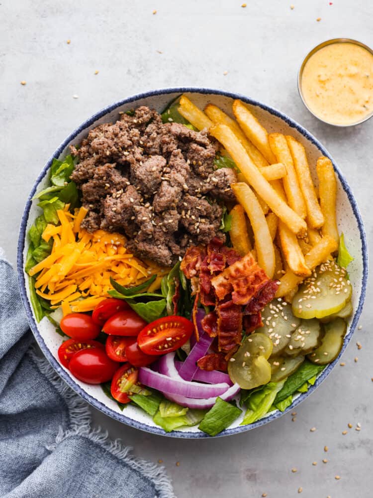 This deconstructed burger bowl is so hearty and satisfying, you'll love every bite! It features crispy fries, tender beef, pickles, and other fresh veggies all coated in homemade Big Mac sauce! Top-down view of a blue and white bowl filled with ground beef, onions, pickles, tomatoes, lettuce, French fries, and bacon, topped with Big Mac sauce.