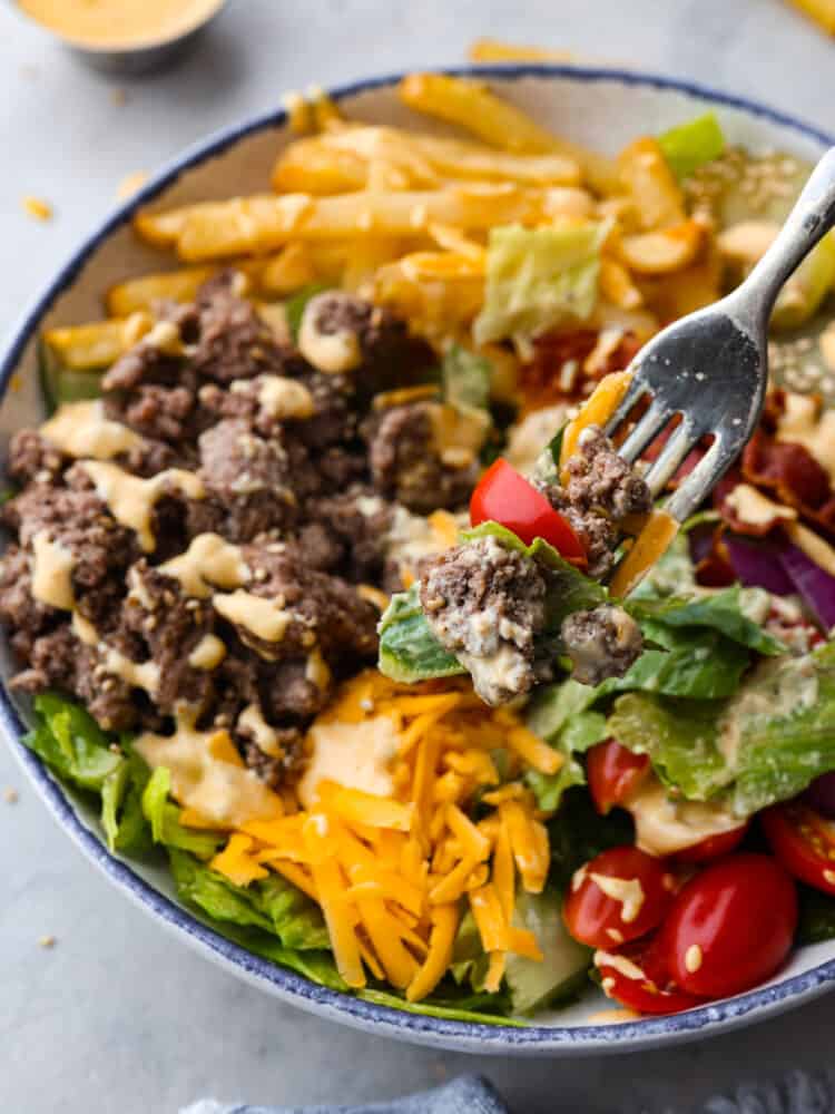 This deconstructed burger bowl is so hearty and satisfying, you'll love every bite! It features crispy fries, tender beef, pickles, and other fresh veggies all coated in homemade Big Mac sauce! Closeup of lettuce, a halved tomato, and ground beef on a fork.