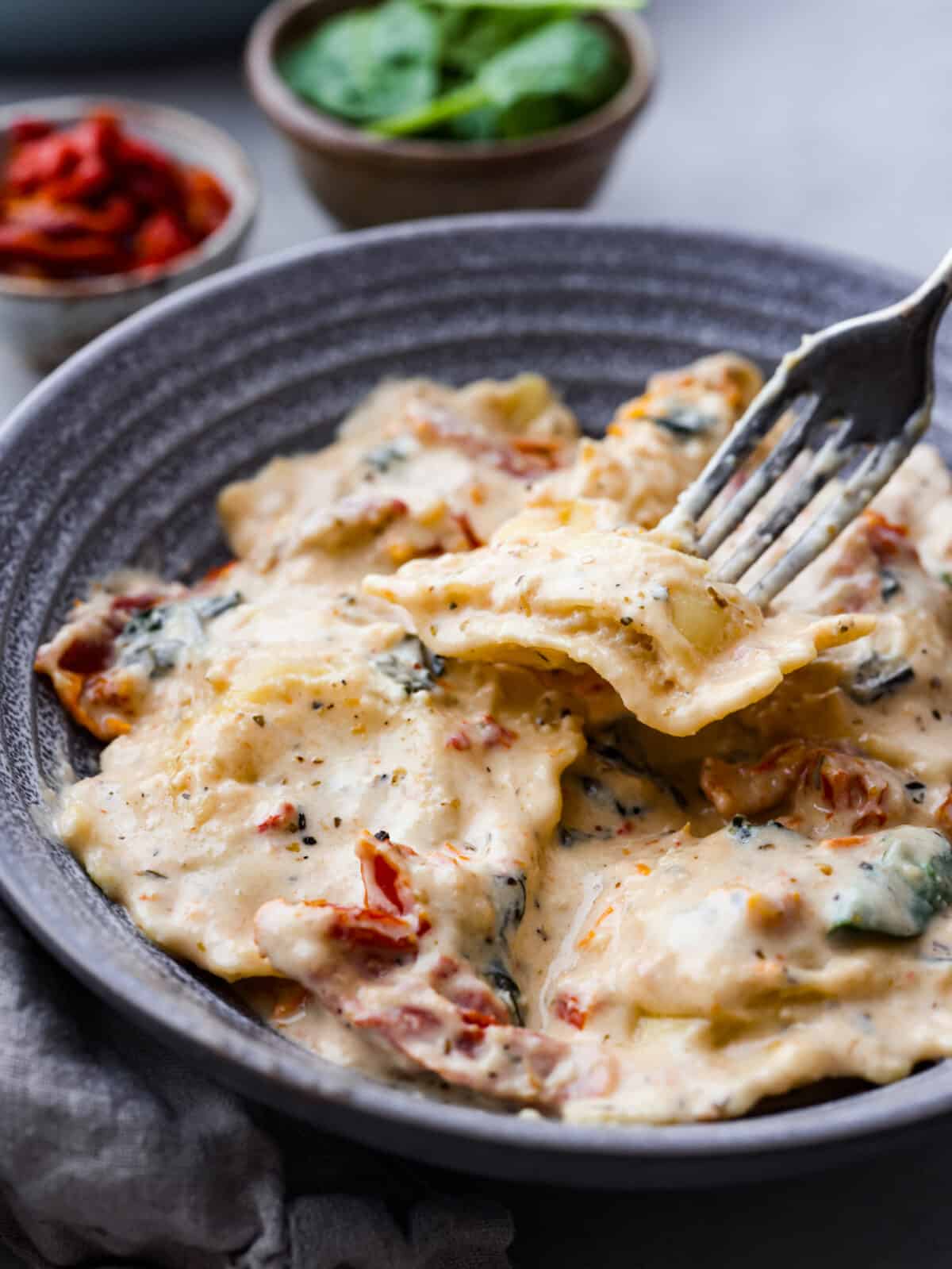 Indulge in this creamy Tuscan ravioli made with the most mouthwatering sun-dried tomato sauce! It's one of the best pasta dishes you'll make, and it's ready in less than 30 minutes. The perfect dinner for busy weeknights! Closeup of ravioli served in a gray stoneware bowl.